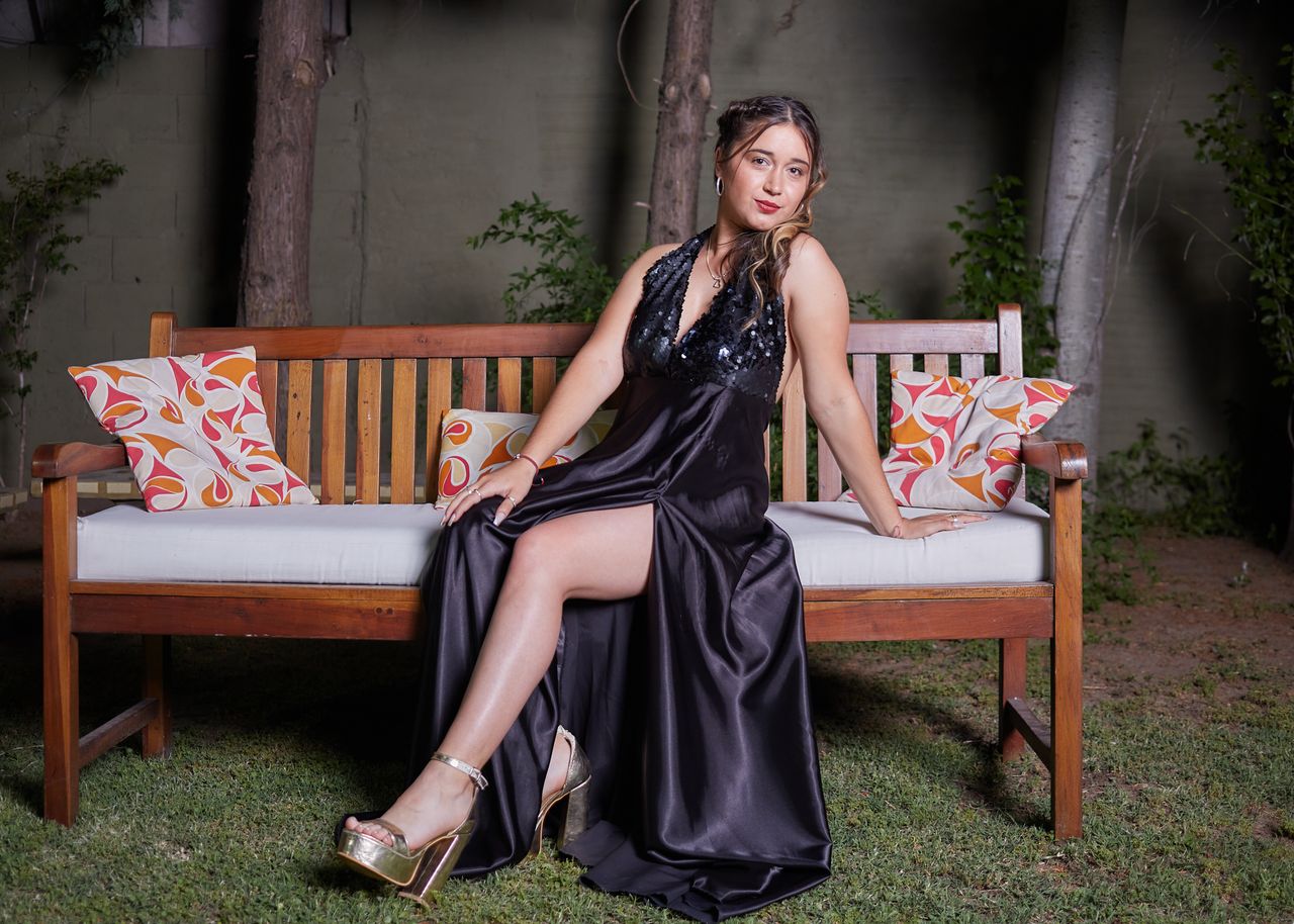 Elegant woman in a black evening dress posing on a bench outdoors, showcasing fashion and style.