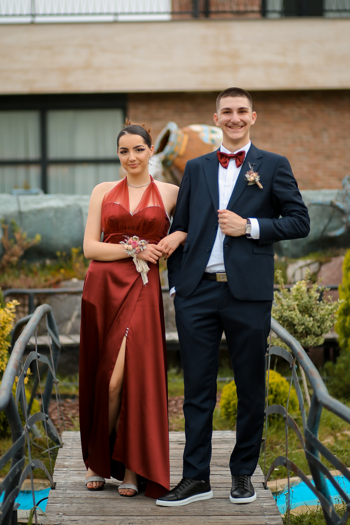 A well-dressed couple stands confidently on a footbridge, showcasing formalwear in an outdoor setting.