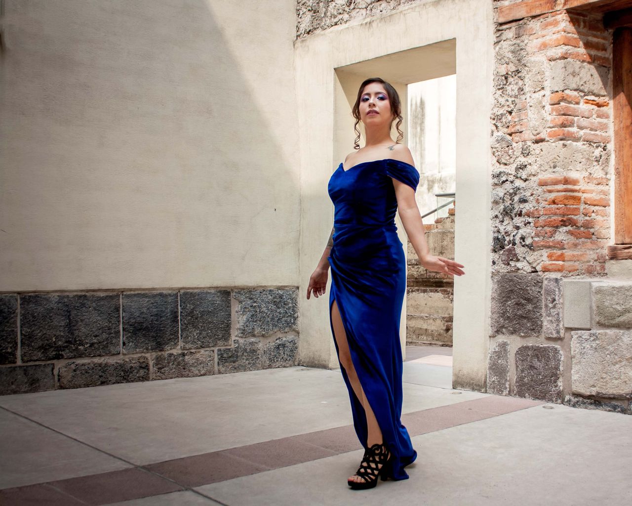 Graceful woman in a blue dress poses against rustic architecture, showcasing elegance and style.